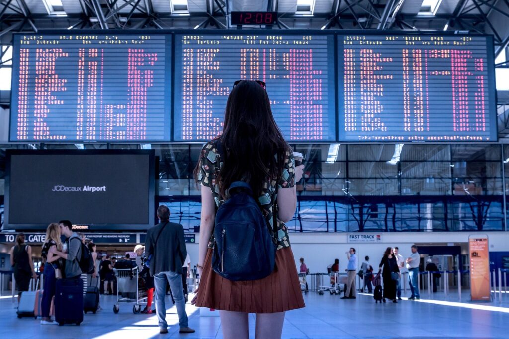 A woman with a backpack stands in front of a large airport departure board, holding a drink and looking up at the flight information.